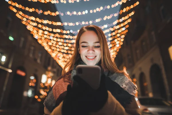 Улыбающаяся девушка использует смартфон на фоне улицы, украшенный рождественскими огнями, смотрит на экран смартфона и улыбается. Рождественский портрет . — стоковое фото