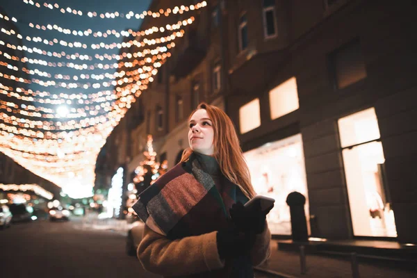 Street night portrait of a cute girl in warm clothes walking down the street with lights, looking to the side and holding a smartphone in her hand. Girl stands on the street decorated with Christmas l — ストック写真