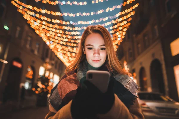 Симпатичная девушка в теплой одежде стоит на фоне рождественских огней и использует смартфон с серьезным лицом. Ночной портрет девушки на улице с смартфоном . — стоковое фото