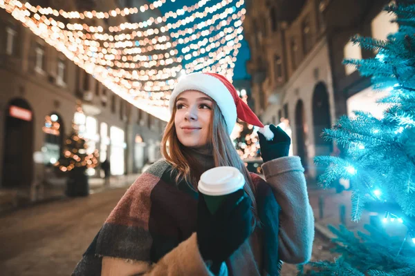 Ελκυστικό κορίτσι σε ένα καπέλο Σάντα και ένα φλιτζάνι καφέ στο χέρι της στέκεται στο δρόμο διακοσμημένο για τα Χριστούγεννα και το χριστουγεννιάτικο δέντρο, κοιτάζοντας μακριά και χαμογελαστό. κορίτσι Santa περπατώντας κατά μήκος του δρόμου των Χριστουγέννων — Φωτογραφία Αρχείου