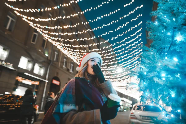 Menina feliz em um chapéu de Papai Noel caminha pela rua no Natal com uma xícara de café em suas mãos, olha para a árvore de Natal e sorri. — Fotografia de Stock