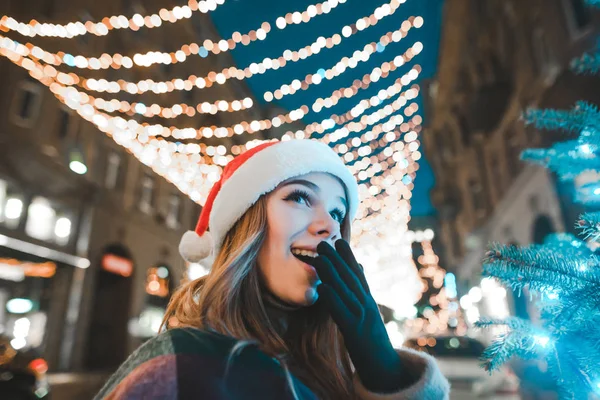 Surprised girl in a hat of Santa Claus looks up at the Christmas tree, stands on a street background with Christmas lights. Evening christmas portrait of shocked cute girl. New Year. — ストック写真