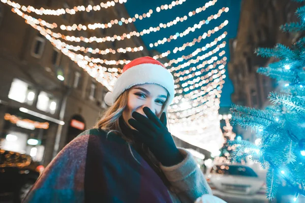 Χαμογελαστό κορίτσι με καπέλο santa στέκεται στο παρασκήνιο του δρόμου διακοσμημένο για τα Χριστούγεννα, χαμογελώντας και κοιτάζοντας την κάμερα. Χαρούμενο κορίτσι, Χριστουγεννιάτικο πορτραίτο. — Φωτογραφία Αρχείου