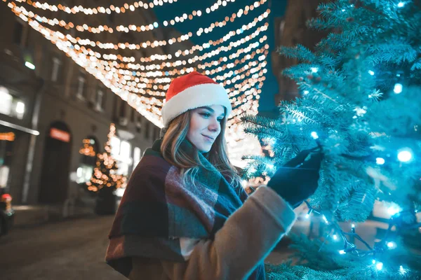 Ελκυστική νεαρή κοπέλα σε ένα χριστουγεννιάτικο καπέλο στέκεται σε ένα διακοσμητικό δρόμο κοντά σε ένα χριστουγεννιάτικο δέντρο και αγγίζει ένα κλαδί. Χαριτωμένο κορίτσι σε Σάντα καπέλο κοντά στο χριστουγεννιάτικο δέντρο με μπλε γιρλάντα. — Φωτογραφία Αρχείου