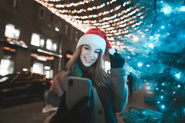 Ευτυχισμένο κορίτσι στο καπέλο Σάντα ποζάρουν σε κάμερα smartphone, κάνει φωτογραφία στο παρασκήνιο των όμορφων Χριστούγεννα διακοσμημένο δρόμο. Χαμογελαστή κυρία λαμβάνοντας selfie στο φόντο χριστουγεννιάτικο δέντρο με μπλε φως — Φωτογραφία Αρχείου