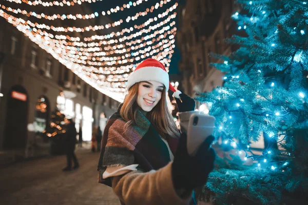 Ένα χαμογελαστό κορίτσι με το καπέλο του Άη Βασίλη στέκεται κοντά σε ένα χριστουγεννιάτικο δέντρο στη διακόσμηση της Πρωτοχρονιάς, βγάζει μια σέλφι σε ένα έξυπνο τηλέφωνο και χαμογελάει. Όμορφη κυρία που βγάζει σέλφι κοντά στο χριστουγεννιάτικο δέντρο. — Φωτογραφία Αρχείου