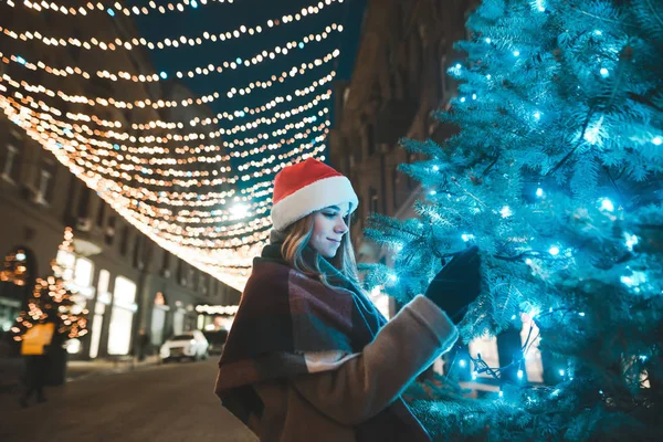 Πορτρέτο ενός όμορφου κοριτσιού σε ένα καπέλο Σάντα στέκεται σε ένα διακοσμημένο δρόμο με μια γιρλάντα και αγγίζει τα κλαδιά ενός χριστουγεννιάτικου δέντρου. Πρωτοχρονιά. Χριστουγεννιάτικο πορτρέτο — Φωτογραφία Αρχείου