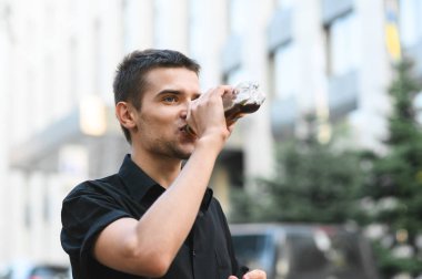 Koyu renk gömlekli genç adamın portresi. Arka plandaki şişeden tatlı su içiyor. Sokak fotoğrafını kapatıyorlar. Yakışıklı genç adam sokakta yürürken karbonatlı bir içki içiyor..