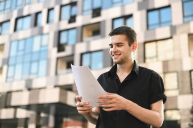 Şehir arka planında elinde belgelerle duran, gazete okuyan ve gülümseyen genç ofis çalışanının portresi. Koyu renk gömlekli meşgul bir adam sokakta yürüyor ve gazete okuyor..