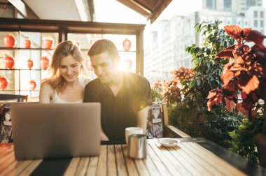 Günbatımı arka planında terasta rahat bir kafede otururken dizüstü bilgisayara bakıp gülümseyen mutlu genç çift. Masadaki restoranda dizüstü bilgisayar kullanan pozitif gençler..