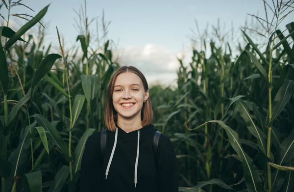 Happy girl stands on a corn field background, looks into the camera and smiles on a garden background with corn. Woman at the cottage standing in the garden growing corn. — Stock Photo, Image
