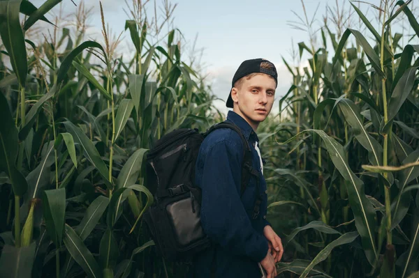 Hombre joven en ropa casual casual y una mochila de pie en un campo con maíz en el fondo, mirando a la camera.Guy en el jardín en una casa de campo sobre un fondo de maíz verde —  Fotos de Stock