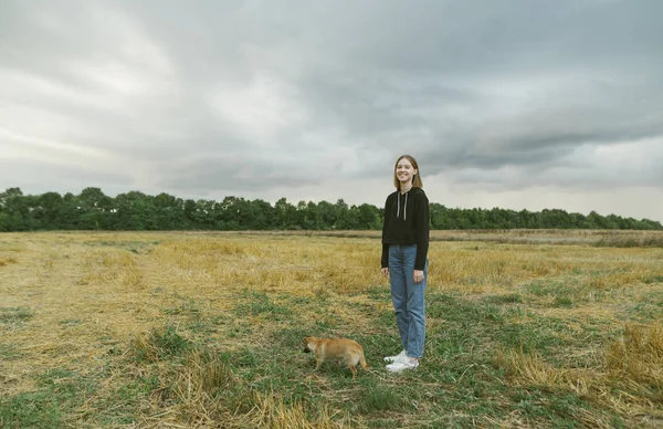 Foto de cuerpo entero del dueño alegre y perrito en el campo de pie sobre la hierba contra el fondo de la naturaleza y el cielo tormentoso, chica mirando a la cámara y sonriendo. Paseando con el perro en el campo . —  Fotos de Stock