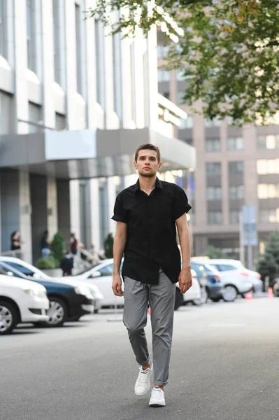 Полный портрет молодого человека в стильной повседневной одежде, в черной рубашке и серых брюках, идущего по городской улице и смотрящего в камеру. Вертикальное фото . — стоковое фото