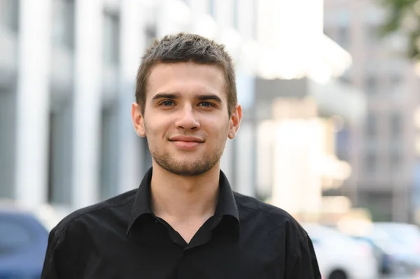 Feliz joven con camisa negra sobre fondo urbano mirando a la cámara y sonriendo. Primer plano retrato de chico guapo en camisa oscura sobre fondo urbano claro . — Foto de Stock