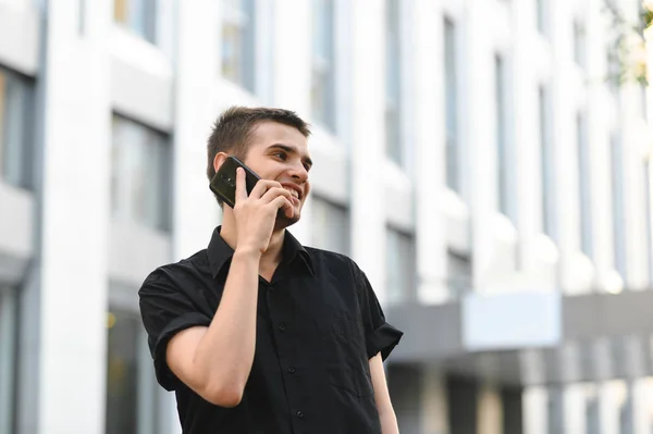 Щасливий молодий чоловік в чорній сорочці перевертає телефон на тлі світлого фону сучасної міської архітектури, дивиться вбік і посміхається. Позитивний бізнесмен розмовляє по телефону . — стокове фото