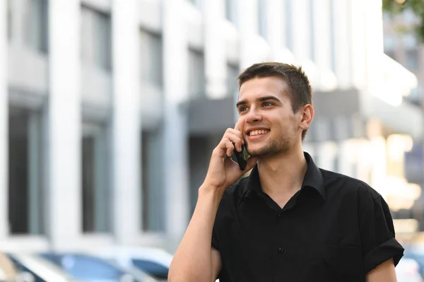 Cheerful young business man in black shirt standing on light urban background and talking on the phone. Portrait of a stylish guy in a shirt uses the phone and smiles.