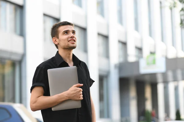 Щасливий молодий бізнесмен з ноутбуком в руці стоїть на легкому міському тлі, посміхаючись і озираючись. Портрет стильного молодого чоловіка, що йде по вулиці з ноутбуком в руці — стокове фото