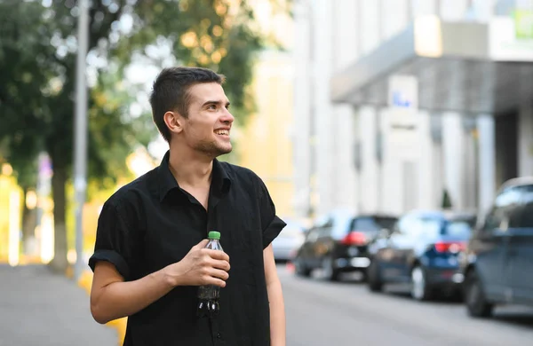 Счастливый молодой человек с темными волосами и стильной одеждой стоит на улице с бутылкой напитка в руке, смотрит в сторону и улыбается. — стоковое фото