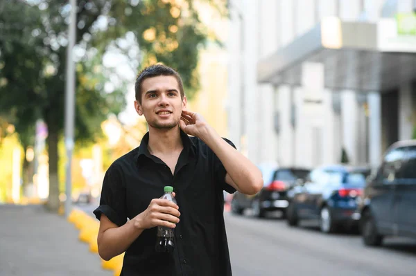 Vonzó fickó sötét ingben és egy üveg édes vízben áll odakint városi háttérben, mosolyog és pózol a kamerába. Stílusos fiatal férfi modell sétál az utcán. — Stock Fotó