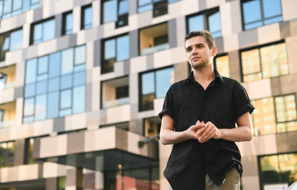 Retrato de un joven guapo con una camisa negra se levanta sobre el telón de fondo de la arquitectura moderna y posa a la cámara, mirando hacia otro lado y sonriendo. Retrato callejero . — Foto de Stock