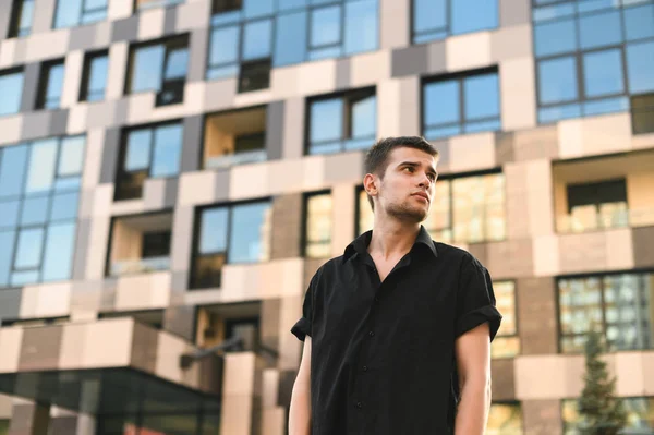 严肃的年轻商人站在城市背景。 穿着黑色衬衫的男模站在一座现代建筑的后面，严肃地把目光移开. — 图库照片