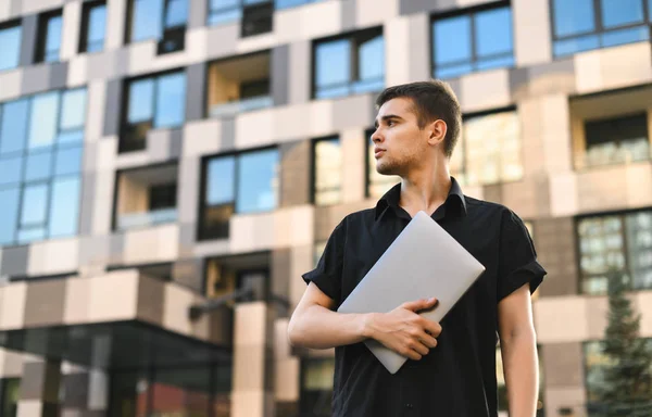 Портрет красивого мужчины в черной рубашке с ноутбуком на фоне современного здания, с серьезным лицом. Студент с ноутбуком стоит на открытом воздухе на современной архитектуре — стоковое фото