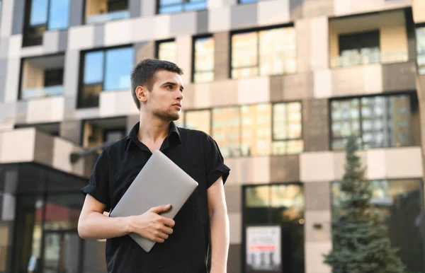 Красивый молодой человек в стильной повседневной одежде стоит на улице с ноутбуком в руке, отворачиваясь, на фоне современной архитектуры. Уличное фото молодого офисного работника с ноутбуком — стоковое фото