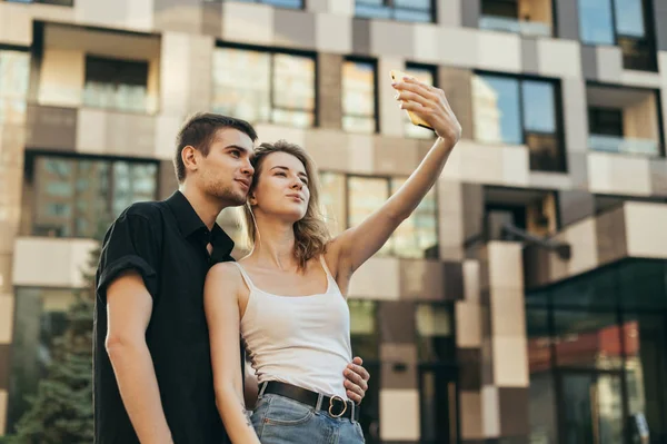 Mooi jong stel staat buiten op een zomerdag en neemt selfie op een smartphone, kijkt naar de camera en glimlacht, draagt stijlvolle casual kleding. — Stockfoto