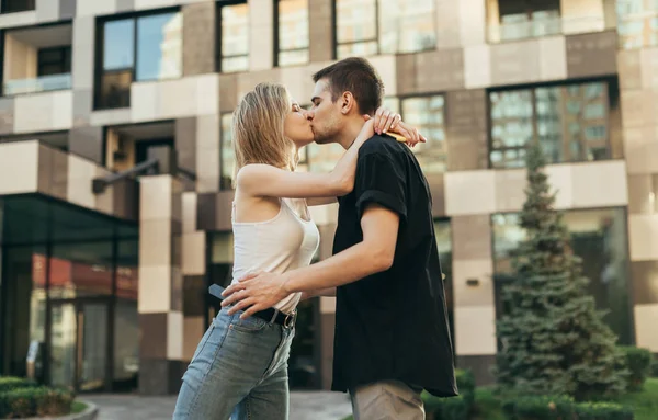Loving young couple in stylish casual clothes kissing outdoors on the background of a modern building. Guy kisses a girl on a walk, she hugs him. Love story concept. — Stock Photo, Image