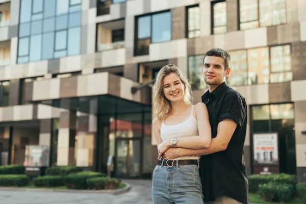 Foto de rua belo casal jovem elegante abraçando contra o pano de fundo de um edifício moderno, olhando para a câmera e sorrindo. Feliz jovem família fica no fundo de um moderno edifício de apartamentos — Fotografia de Stock