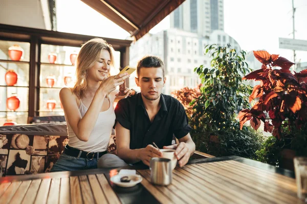 Vackra unga par sitter på terrassen på restaurang vid solnedgången och dricka kaffe, glad flicka tar foto på smartphone kamera och ler. Ung man och flicka sitter vid bordet på caféet. — Stockfoto