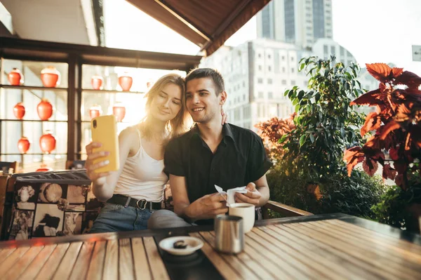 Счастливая любящая пара делает селфи в кафе за столом на фоне заката. Симпатичная улыбающаяся пара, сидящая в уюте. ресторан на террасе и фотографирование на камеру смартфона . — стоковое фото
