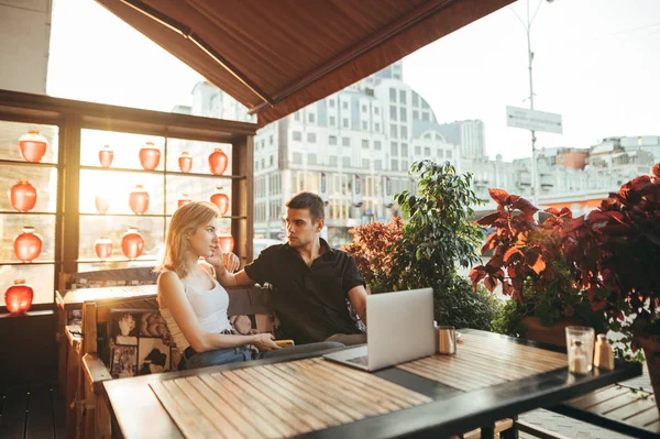 Porträtt av glada kärleksfulla par sitter i mysiga café på terrassen på kvällen och med hjälp av laptop, flicka med smartphone i handen tittar på pojkvän. Några frilansare som jobbar på kafé. — Stockfoto