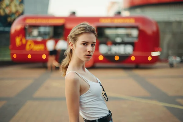 Menina bonita em roupas casuais fica no fundo urbano, olha para a câmera com um rosto sério. Mulher de t-shirt branca posando no fundo Food truck — Fotografia de Stock