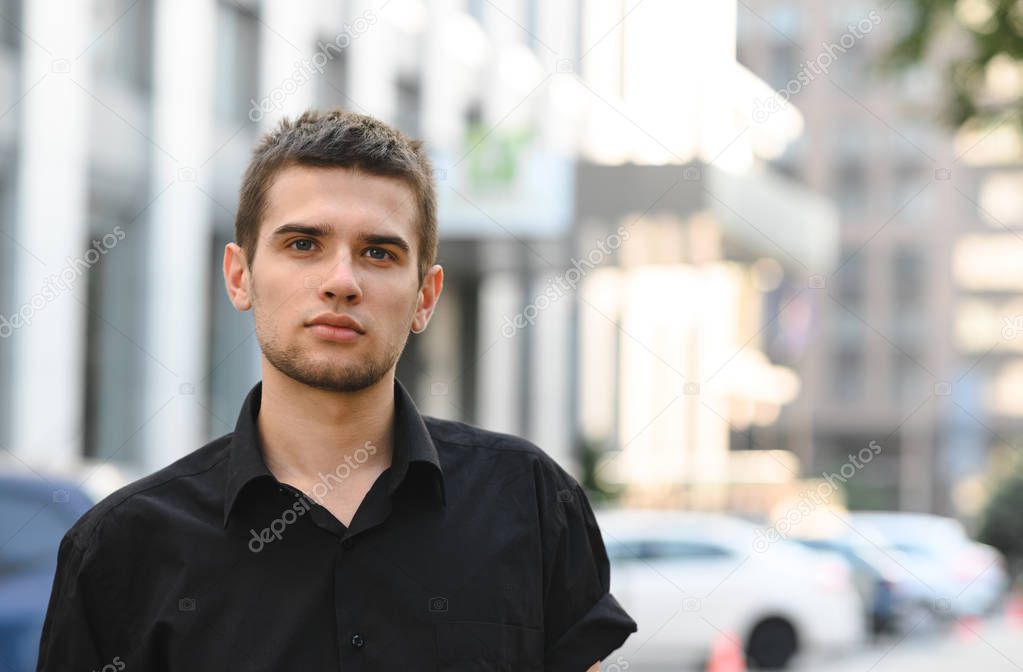 Closeup portrait of a handsome young man in a black shirt on a light cityscape background, looking into the camera with a serious face. Portrait of a guy in a dark shirt.