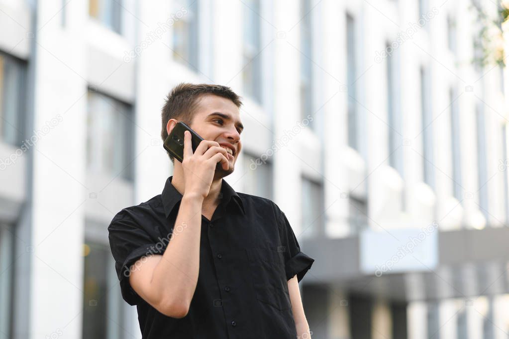 Happy young man in black shirt flips over phone against light background of modern urban architecture, looks away and smiles. Positive business man talking on the phone.