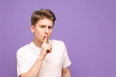 Beyaz tişörtlü adam parmağını dudaklarına koydu ve mor bir arka planda izole edilmiş sessizlik işareti gösterdi. Sessiz olun, lütfen diyor. Ciddi genç adam konuşmamak istiyor, bir jest.