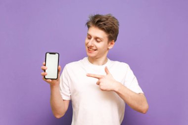 Elinde akıllı telefon olan, beyaz tişört giyen beyaz bir ekranda parmağını gösteren ve mor arka planda izole edilmiş bir adamın portresi. Kopyalama alanı