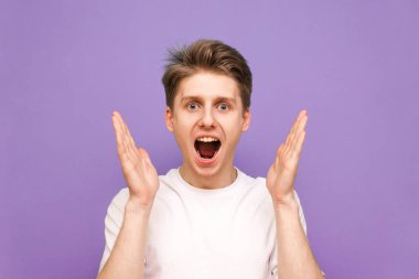 Ağzı açık, kameraya bakan, mor arka planda izole edilmiş beyaz bir tişört giyen şaşırmış bir çocuğun portresini kaybettiler. Şok olmuş bir adam, elleri yüzünün yanında kameraya bakıyor..