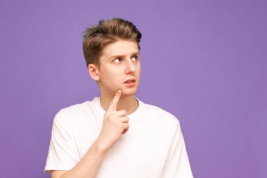 Portrait of a guy in a white T-shirt stands on a purple background, looks aside and thinks. Thoughtful young man poses on a blue background and looks at a place for advertising. Isolated.