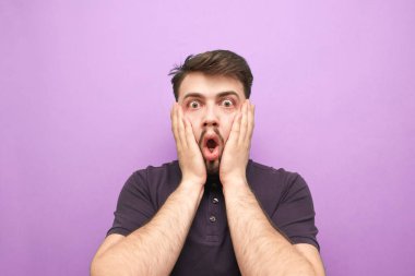 Korkunun yüzüne dokunan şaşırmış bir adamın yakın plan portresi. Mor arka plandaki kameraya duygusal olarak bakıyor. Mor bir duvarın arkasında sakallı, şok olmuş bir adam.