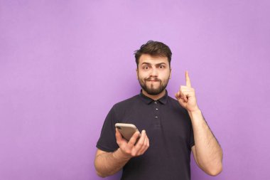 Mor arka planda elinde telefon olan sakallı komik bir adamın portresi kameraya bakıyor ve komik bir yüz ifadesi takınıyor. Akıllı telefonlu kafası karışmış bir adam boş bir alan gösteriyor. İzole edilmiş.