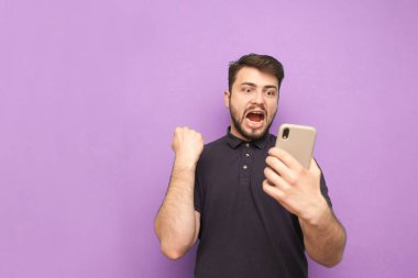 Pozitif genç adam elinde bir akıllı telefon tutuyor, ekrana bakıyor ve elleri havada seviniyor. Mor bir arka planda izole edilmiş bir şekilde gülümseyen ve sevinen adam. Kopyalama alanı