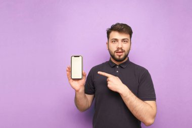 Sakallı, koyu renk tişörtlü, elinde beyaz ekranlı akıllı bir telefon tutan, parmağını gösteren, mor arka plandaki kameraya bakan duygusal bir adam. Yetişkin adam akıllı telefon ekranında görünüyor.