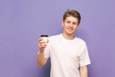 Beyaz tişörtlü mutlu genç adam elinde mor menekşe rengi bir fincan kahve tutuyor.
