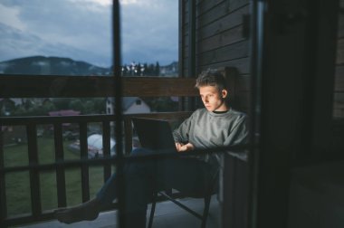 Konsantre olmuş genç bir adam bir apartmanın balkonunda bir sandalyede oturuyor ve dizüstü bilgisayar kullanıyor, dikkatle bilgisayar ekranına bakıyor. Freelancer balkonda dizüstü bilgisayarda çalışıyor..