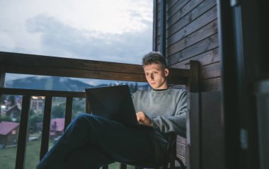 Laptopta çalışan yakışıklı bir adam, akşam otelin balkonunda oturuyor, dizüstü bilgisayarın ekranına bakıyor. Dairede dizüstü bilgisayar kullanan genç bir adamın portresi güzel manzaralı.