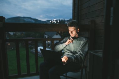 Düşünceli genç adam akşamları bir apartmanın balkonunda bir sandalyede oturur ve ciddi bir yüzle ekranda dizüstü bilgisayar kullanır. Akşam evde dizüstü bilgisayarla çalışıyorum..