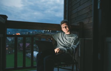 Kucağında dizüstü bilgisayarla balkonda oturan mutlu bir gencin portresi. Güzel manzaralı dairelerde dizüstü bilgisayar kullanan bir adam.